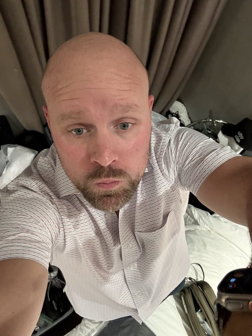 A selfie of a bald man with a beard wearing a light grey short-sleeved button-up shirt, sitting on a bed in what appears to be a hotel room.