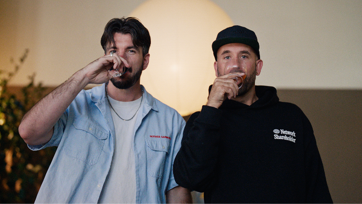 Two men drink from small glasses in front of a glowing round light, with one wearing a light blue shirt and the other in a black "Newport Skateboards" hoodie, playfully illustrating what Square had to say.