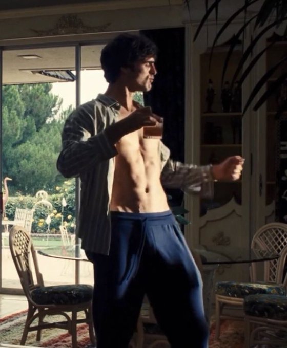 The image features a shirtless man standing in front of a window, holding a glass of wine or beer while wearing blue sweatpants. The background includes chairs and a dining table, suggesting that he might be in a living room setting. The caption "the kind of guy you pull when mega eth finally cooks" implies that the man is likely a famous actor or someone with significant influence.