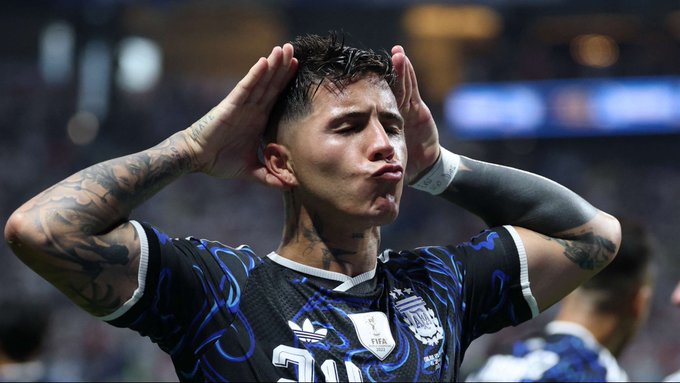 Argentine soccer player Enzo Fernández in an Argentina national team jersey, making a celebratory gesture with his hands near his head, featuring visible arm tattoos.