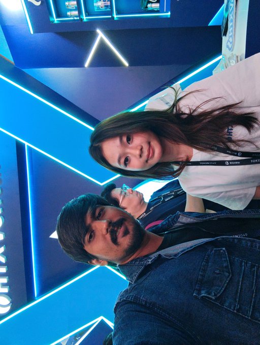 A sideways selfie shows a smiling woman in a white top with a 'TOKEN 2049' lanyard and a man with a mustache in a denim jacket, standing in a venue with blue neon lighting.