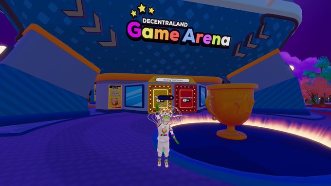 A screenshot from the virtual world Decentraland showing the 'Game Arena' with an avatar standing in front of colorful game machines and a large golden trophy cup.
