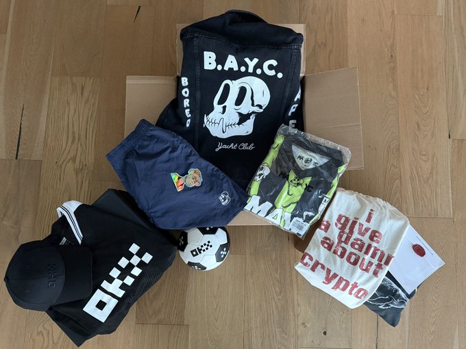A collection of streetwear and accessories including a B.A.Y.C. Bored Ape Yacht Club hoodie, OKX-branded clothing, a soccer ball, and a 'I give a damn about crypto' t-shirt laid out on a wooden floor.
