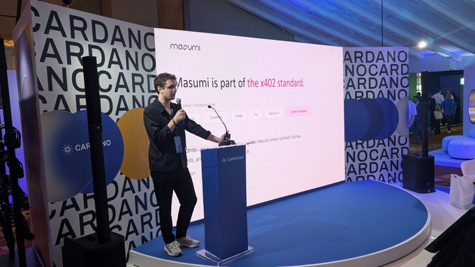 A man stands at a podium on a stage at a Cardano event, presenting a screen that reads 'Masumi is part of the x402 standard.'