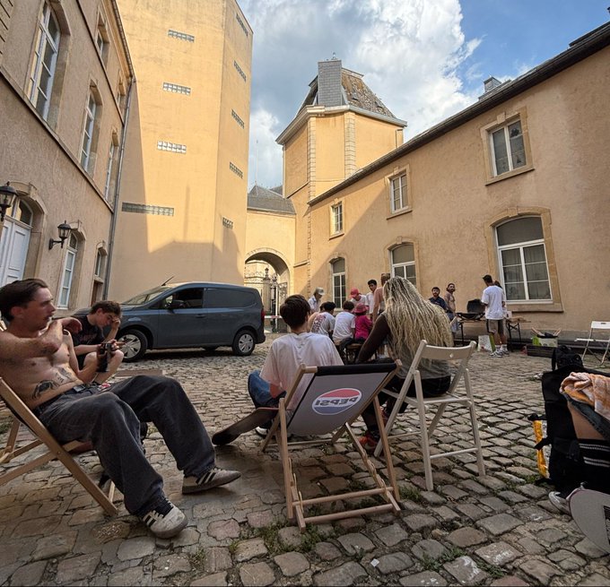 People gather in a cobblestone courtyard surrounded by yellow buildings, sitting on folding chairs while one shirtless man holds a camera.