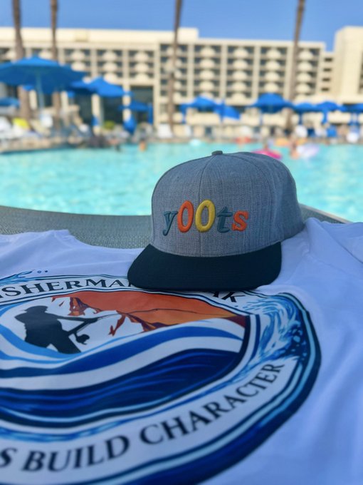 A gray 'Yoots' snapback hat resting on a Sherman Oaks 'Fishermen Build Character' t-shirt, with a resort pool and beach umbrellas visible in the background.