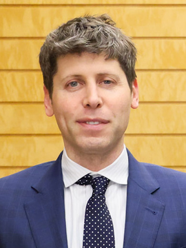 A man with short brown hair wearing a navy blue suit, white shirt, and polka dot tie stands in front of a yellow wooden wall.