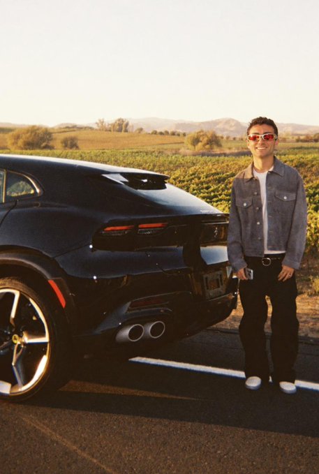 A man wearing red sunglasses stands beside a black Ferrari sports car parked beside a vineyard at sunset. The car’s rear badge and license plate are visible but not clearly readable.
