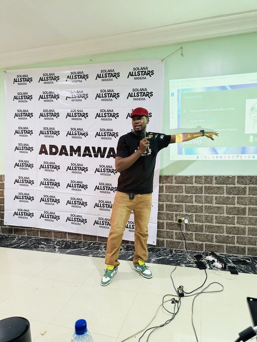 A man wearing a black t-shirt, red cap, and green sneakers speaking into a microphone at a Solana Allstars Nigeria event, with 'ADAMAWA' visible on the branded backdrop.