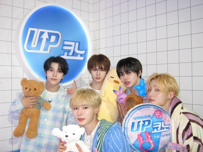 The image features a group of five young men posing together for a photo, with each person holding up a stuffed animal. The stuffed animals include a teddy bear and two other plush toys. In the background, there's a blue sign that reads "UPKONO®". This suggests that they might be at an event or gathering related to UPKONO® or another brand. The image also contains text in Korean, which could indicate that it was taken during a time when the language is spoken or that the group consists of individuals who speak Korean as their first language.