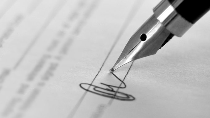 A black-and-white close-up photograph of a fountain pen nib signing a document on lined paper.