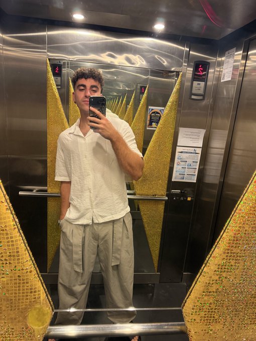 A young man in a cream short-sleeve shirt and beige pleated cargo pants takes a mirror selfie inside a striking gold-tiled elevator, capturing his arrival in Singapore amid the haze of jet lag and humidity for the Solana Summit.