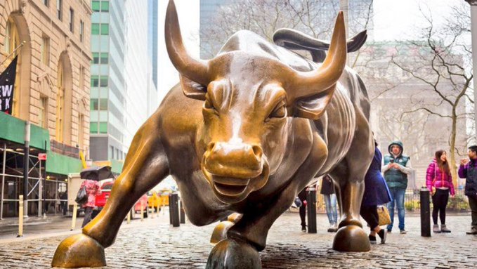 The image shows the iconic Charging Bull statue on Wall Street in New York City, a famous symbol of financial optimism and bullish markets, with pedestrians walking nearby on the cobblestone street. The statue's powerful, forward-charging stance visually reinforces the post's bullish message about a Bulgarian bank opening Bitcoin and crypto access to over 1 million clients, symbolizing a significant stride forward in mainstream crypto adoption.