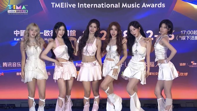 Six young women in coordinated white and pink stage outfits pose together on stage at the "TMElive International Music Awards."