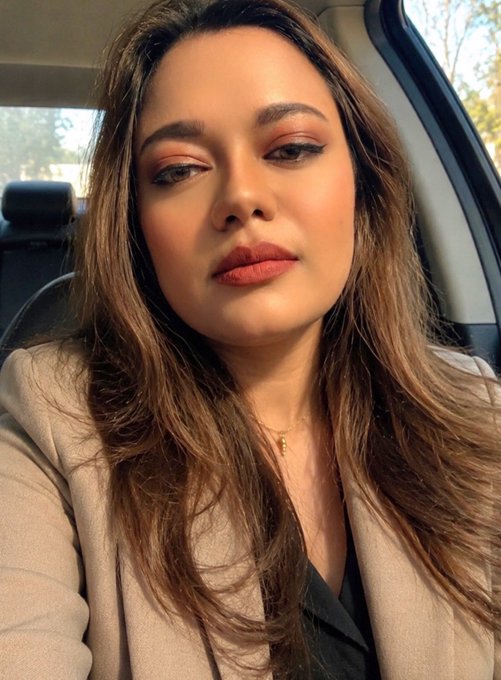 A woman with long brown hair and makeup takes a selfie while sitting inside a car.