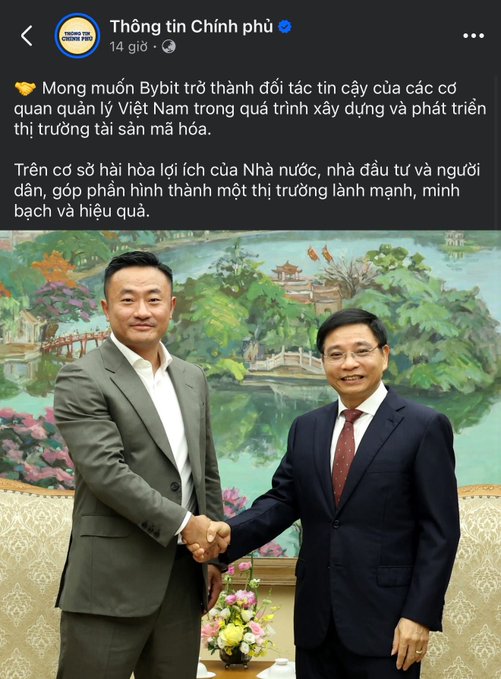 The image shows two men shaking hands over a table with flowers. The man on the left has his hand outstretched towards the other man's hand, while the second man stands behind him. They are both dressed professionally, wearing suits and ties. In the background of the photo, there is a painting depicting a river surrounded by trees, adding an artistic touch to the scene.