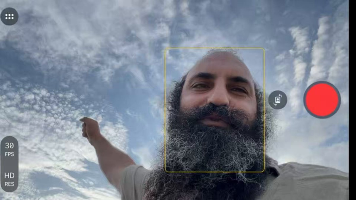 The image shows a man with a beard taking a selfie using his phone's camera. The photo captures him from behind, making him appear larger than life due to the angle of the shot. The background features a blue sky with clouds, providing a beautiful backdrop for the photograph. The image also includes text that reads "BITCOIN TRADE BUILDING, NVIDIA DROP ON REBALANCE AT MARKET CLOSE $BTC $NVDA".