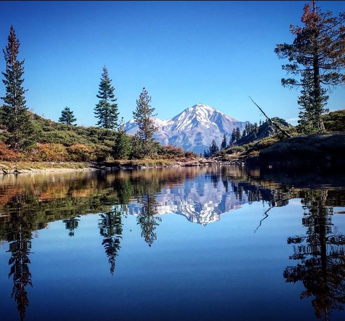A scenic landscape photograph showing a snow-capped mountain reflected in a calm alpine lake, surrounded by evergreen trees under a clear blue sky.