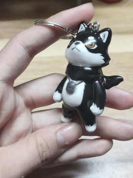 A hand holds a small black and white cat-shaped keychain with a bell collar attached to a silver keyring.