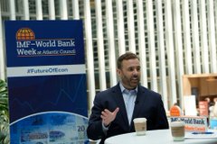 The image features a man wearing a suit, sitting at a table with two cups of coffee placed in front of him. The background includes a white building and a blue sign that reads "IMF-World Bank Wee". The text on the sign suggests that the event is related to the World Bank or the International Monetary Fund.