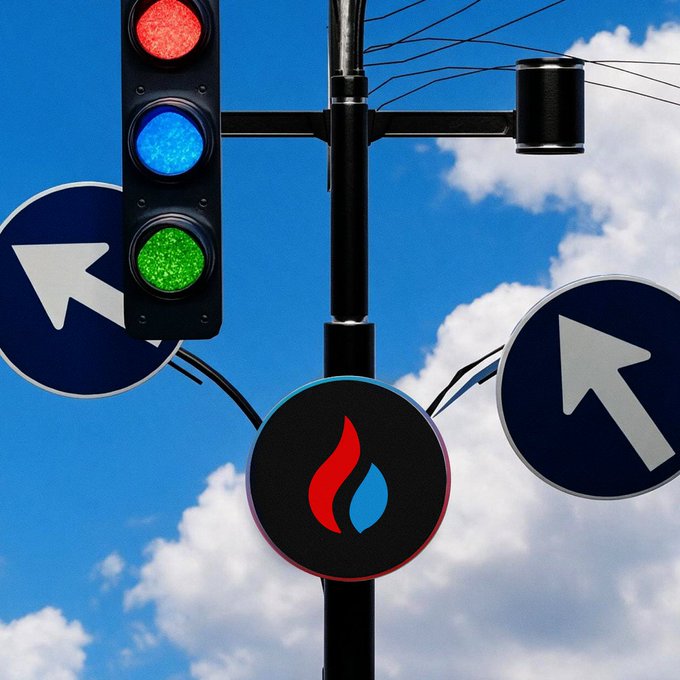 A traffic light with red, blue, and green signals mounted on a pole with directional arrow signs and a fuel/gas station logo against a blue sky with clouds.