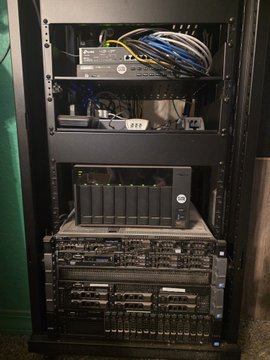 A black server rack containing networking equipment, including a switch with multiple ethernet cables plugged in, and several rack-mounted servers below it.