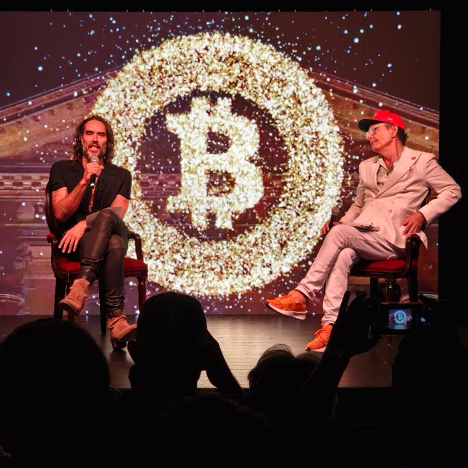 Two men sit on stage in front of a large Bitcoin logo made of glittering gold sparkles at the "BITCOIN HISTÓRICO" conference held November 11-12 at the National Palace. One speaker holds a microphone while discussing the conference's theme of "Life. Liberty. Bitcoin," with audience members visible in the foreground watching the panel.