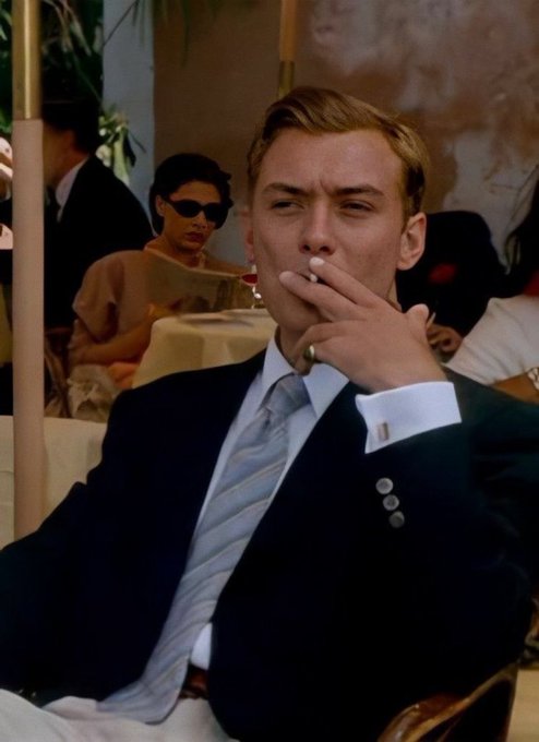 A scene from a film featuring a blond man in a dark suit and striped tie smoking a cigarette, with other people in the background.