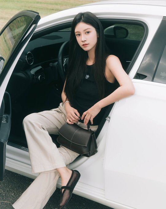 A young woman with long black hair wearing a black tank top and beige wide-leg pants sits in the open door of a white car, holding a black handbag, with a grassy field in the background.