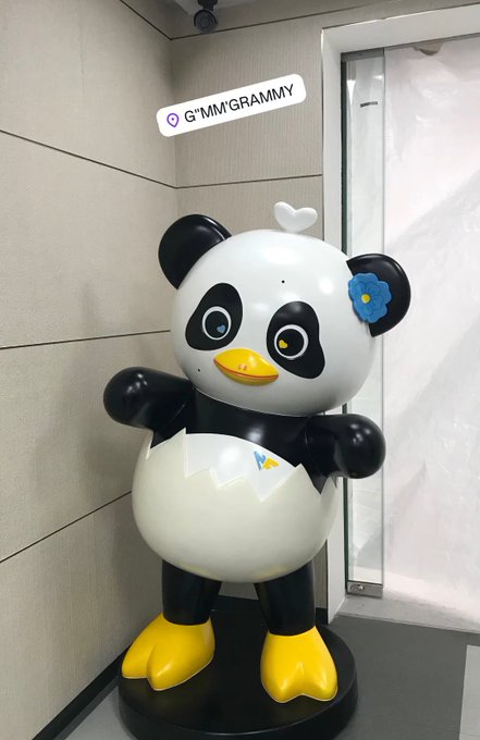 A cute cartoon panda mascot stands on a black pedestal, emerging from a cracked eggshell with a yellow beak and yellow feet, decorated with a small blue flower and white heart on its head. The text bubble reads "📍G'MMGRAMMY," fitting the post's "#LUNAR" theme as the adorable mascot appears to be debuting or hatching at the location, likely a promotional event.
