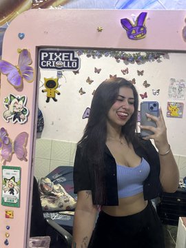 A mirror selfie of a smiling young woman with long dark hair, wearing a blue top and black cardigan, holding a smartphone, with a decorated mirror frame featuring butterfly and character stickers, one reading 'PIXEL WORLD'.