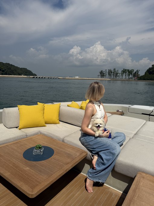 A woman with short hair sitting on a luxury boat's outdoor lounge area with yellow cushions, holding a small white dog in her lap, looking out at the water and distant shore.