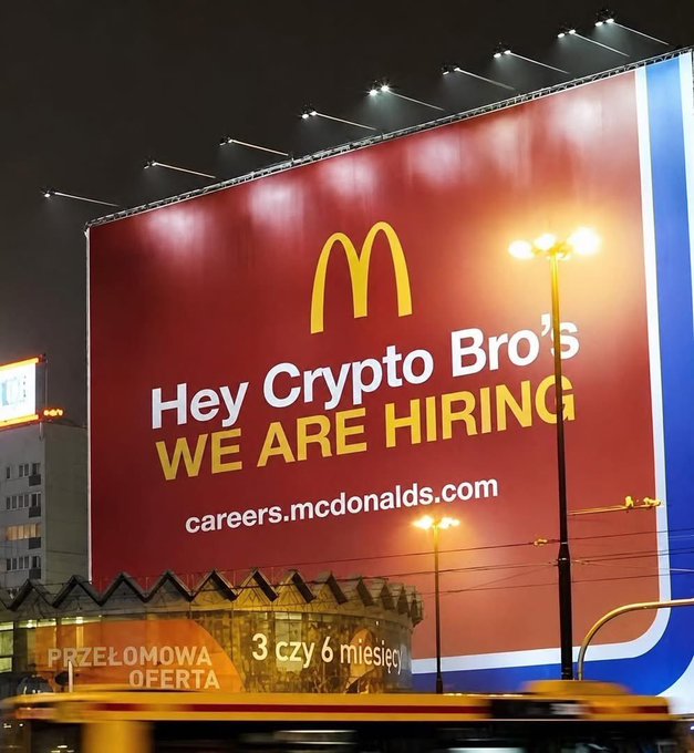 The image features a large billboard with a red background, white text, and yellow text that reads "hey crypto bros we are hiring". The billboard also includes an advertisement for McDonald's restaurants. In the foreground of the image, there is a bus driving by on the street below the billboard.