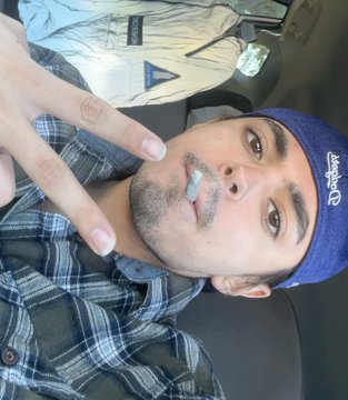 A selfie shows a bearded man wearing a blue bandana and plaid shirt making a peace sign, photographed from an overhead angle.