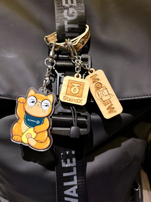Photograph of three keychain charms attached to a black bag strap: an orange cartoon cat with 'KITTAYAMA' on its shirt, a wooden 'StandX' tag, and a wooden 'MOE M EW' tag.