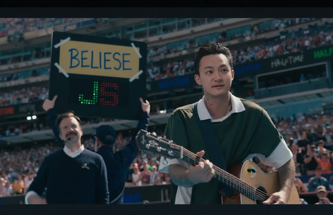 An Asian man playing an acoustic guitar at what appears to be a baseball stadium (Yankee Stadium visible), with a sign reading 'BELIESE' held up in the crowd behind him.