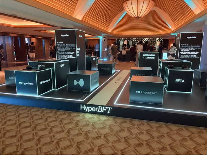 A Hyperliquid ("HyperBFT") event booth displays illuminated cubes featuring logos for Stablecoins, Spot, Oracles, Bridging, NFTs, and Liquid, with attendees gathered in the venue—setting the scene for the chaotic college kids buying merch for "5 hype" and drunkenly declaring loyalty to "Jeff."