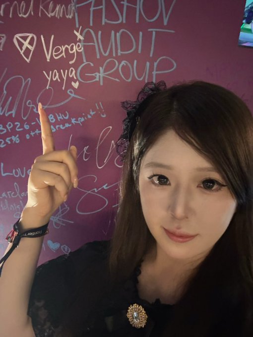 A young woman wearing a black lace headband and dark outfit pointing upward, posed in front of a pink wall covered in signatures and messages including "Verge" "FASHION AUDIT" and "yaya GROUP."