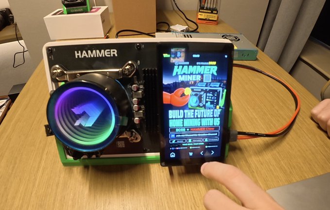 A custom PC build labeled 'HAMMER MINER' with RGB cooling fan and an attached screen displaying mining software, sitting on a wooden desk.