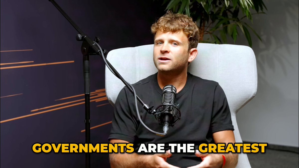 The image features a man sitting in a chair, wearing a black shirt with white text that says "Governments are the greatest". The background shows a microphone and speaker setup, suggesting this could be part of a podcast or radio show.