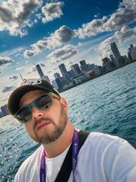 A selfie of a man wearing a cap and sunglasses, with a city skyline visible across a body of water in the background.