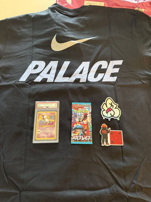 A black Palace Nike t-shirt laid flat, displaying the Nike swoosh and 'PALACE' in white, with several items on top including a graded Pokémon Charizard card, an 'Our War' trading card, and two character patches/stickers.