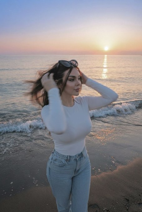A young woman in a white long-sleeve top and light blue jeans posing on a beach during a scenic sunset, with sunglasses resting on her head.