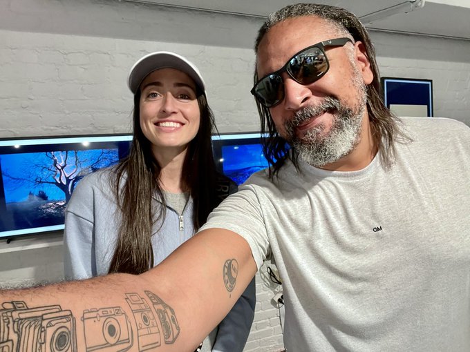A selfie of a smiling woman wearing a white cap and a man with dreadlocks and sunglasses, with the man's tattooed arm showing camera illustrations in the foreground.