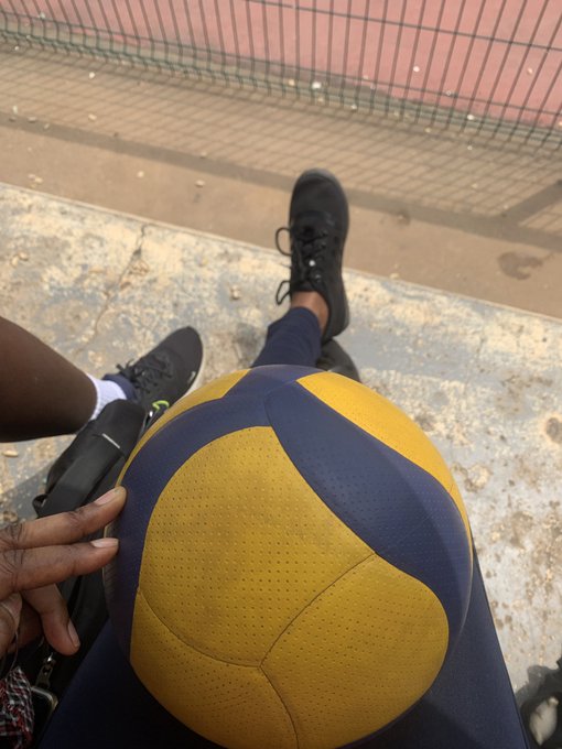 A first-person perspective photo showing a yellow and blue volleyball held between the photographer's legs, with another person's foot wearing a black sneaker visible on a concrete surface next to a chain-link fence.