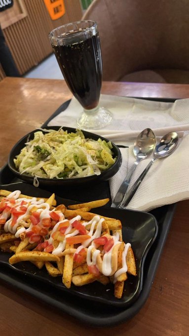 A photo of a meal on a black tray featuring a plate of fries topped with diced tomatoes and white sauce, a side salad with creamy dressing, and a tall glass of dark soda with utensils.