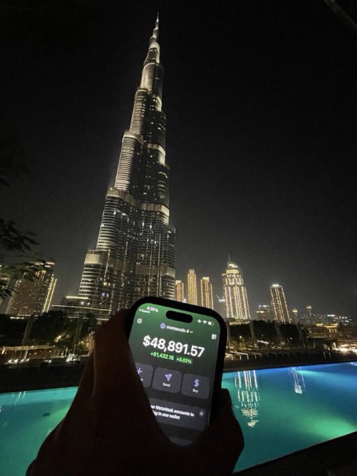 A nighttime view of the Burj Khalifa in Dubai with a hand holding a smartphone in the foreground displaying a wallet balance of $48,891.57.