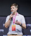 The image features a man standing on a stage with a microphone, wearing a white shirt and khaki pants. He has an orange lanyard around his neck that displays the name "@beyondtechexpo" along with other information about the event. The background of the image is black, which contrasts with the white text displayed in the post. The post mentions various topics such as talks on AI and the trust layer for AI, booth demos, project teams, and builders testing the stack in person. It also includes a link to a video that provides more information about these topics.