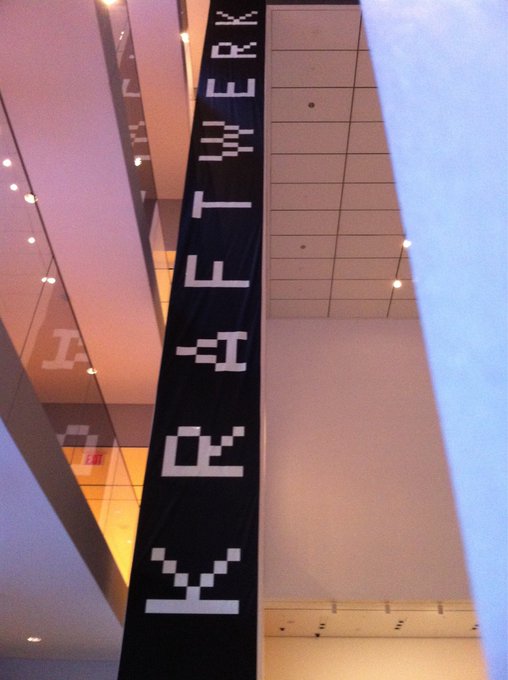 A tall vertical sign displays the pixelated text "KRAFTWERK" in a digital font, set against the modern architectural backdrop of what appears to be a museum or gallery interior. The blocky, computer-style typography reflects the German electronic music group's pioneering aesthetic of machine-like precision and digital-age imagery that influenced the crypto/tech culture referenced in the post.