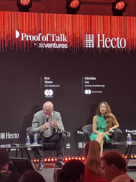 The image shows a man sitting at a table with a microphone while another woman sits next to him. They are both wearing suits and appear to be engaged in conversation, possibly discussing innovation or business strategies. The background features a red curtain that has "proof of talk" written on it along with the words "X Ventures".